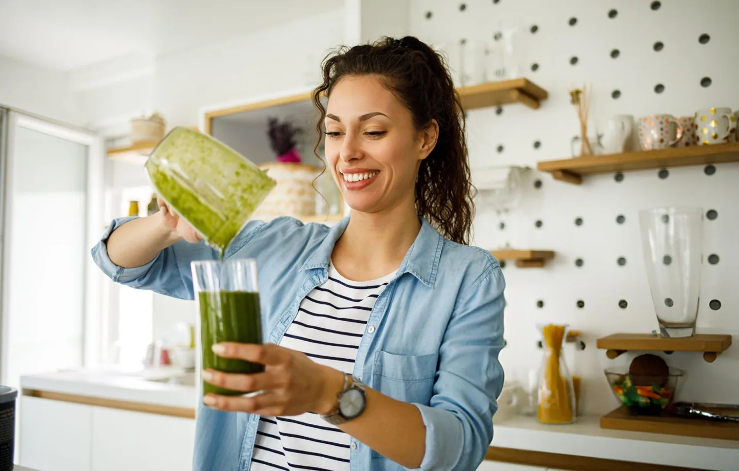 Smiling woman in a modern kitchen pouring a fresh green smoothie from a blender into a glass. She wears a striped shirt and a denim jacket, with wooden shelves and kitchen decor in the background. The scene conveys a healthy lifestyle and homemade detox drinks.