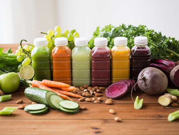 Six bottles of colorful fresh juice, arranged in a row on a wooden table, surrounded by fresh ingredients like kale, carrots, cucumbers, beets, almonds, and grapes. The juices range from green to orange, yellow, red, and purple, symbolizing a variety of detox and health-boosting drinks.