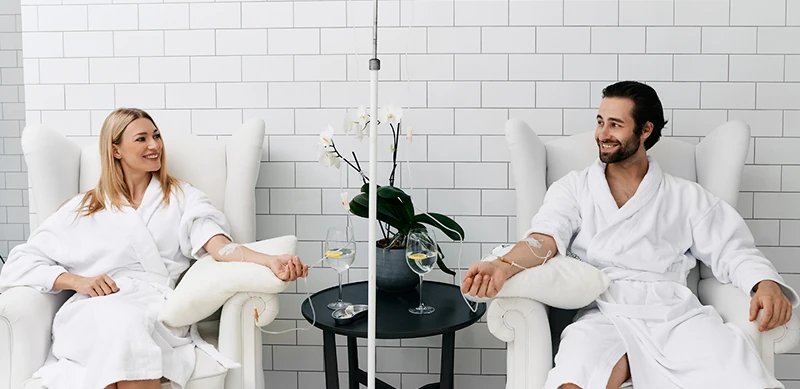 A smiling man and woman in plush white robes relaxing in comfortable chairs while receiving IV therapy. They are holding hands and enjoying glasses of water or a light beverage, creating a luxurious spa-like atmosphere with a serene, rejuvenating ambiance.