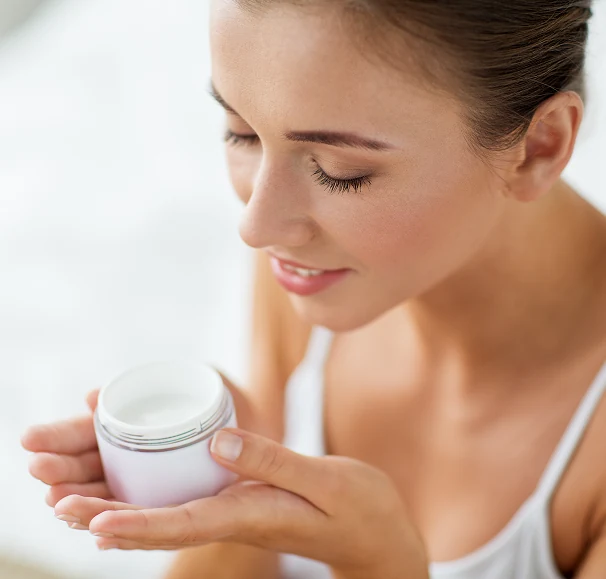 A young woman with glowing skin holding a jar of cream, smiling as she gently smells it. She is wearing a white tank top, and the background is softly blurred, creating a serene and fresh atmosphere.